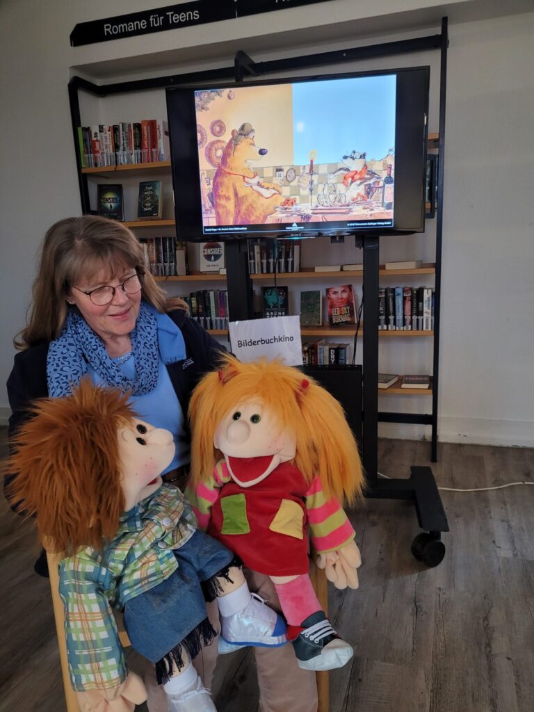 Helga Rebling sitzt mit zwei Handpuppen auf dem Schoß vor einem Monitor, auf dem ein Bild mit einem Bären aus einem Bilderbuch zu sehen ist.