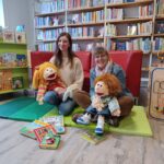 Gina Friedrichs und Helga Rebling sitzen mit den Stoffpuppen Lilabelle und Fabian auf dem Schoß in der Kinderbücherei und lächeln in die Kamera.